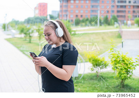A beautiful fat girl in black sportswear listens to music on headphones and text messages on the phone while walking outdoors. A plump young woman is leafing through a music playlist in smartphone. 73965452