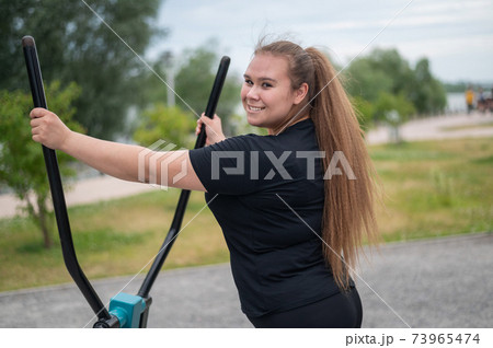 Beautiful smiling overweight young woman does fitness on an ellipsoid simulator outdoors. Fat girl is training on the sports ground for weight loss. Sport outside on a warm summer day. 73965474