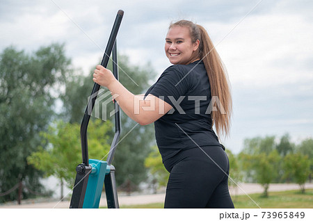 Beautiful smiling overweight young woman does fitness on an ellipsoid simulator outdoors. Fat girl is training on the sports ground for weight loss. Sport outside on a warm summer day. Beautiful smiling overweight young woman does fitness on an ellipsoid simulator outdoors. Fat girl is training on the sports ground for weight loss. Sport outside on a warm summer day. 73965849