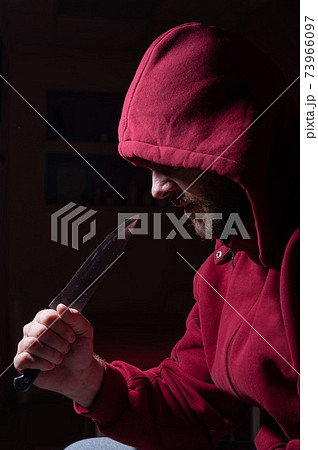 A dangerous bearded man in a red hood holds a sharp knife near his face. A criminal or a killer with knives in the dark. A dangerous bearded man in a red hood holds a sharp knife near his face. A criminal or a killer with knives in the dark. 73966097