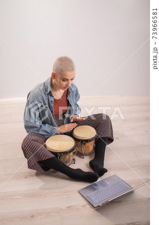 A young hipster woman sits on the floor at home and watches educational videos on mini bongs. Girl remotely learns to play traditional ethnic drums. A young hipster woman sits on the floor at home and watches educational videos on mini bongs. Girl remotely learns to play traditional ethnic drums. 73966581
