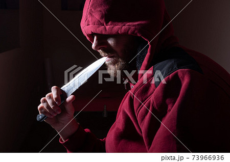 A dangerous bearded man in a red hood holds a sharp knife near his face. A criminal or a killer with knives in the dark. 73966936