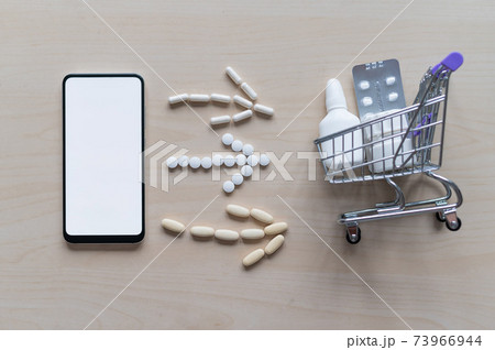Online pharmacy concept with home delivery. Buying medicine online by phone. Mini trolley with different tablets, sprays, capsules and a smartphone with a white blank screen. Arrow made of pills. 73966944