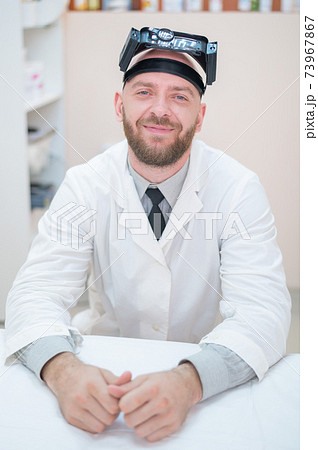 Male bearded doctor in a medical coat and with a magnifying head strap w lights sitting on the desk. Optometrist equipment. Medical office. Male bearded doctor in a medical coat and with a magnifying head strap w lights sitting on the desk. Optometrist equipment. Medical office. 73967867