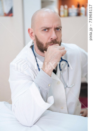 A male doctor sits at a desk in a medical office. A bald man with a beard works as a therapist in a clinic. Serious cardiologist with a stethoscope in a lab coat. Vertical photo. A male doctor sits at a desk in a medical office. A bald man with a beard works as a therapist in a clinic. Serious cardiologist with a stethoscope in a lab coat. Vertical photo. 73968117