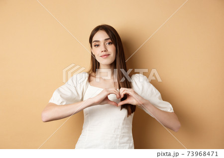 Valentines day and romance concept. Romantic caucasian girl in white dress showing heart gesture and smiling, say I love you, standing over beige background Valentines day and romance concept. Romantic caucasian girl in white dress showing heart gesture and smiling, say I love you, standing over beige background 73968471