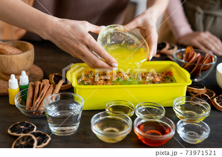Hands of female pouring melted soap mass into container with dry flowers 73971521