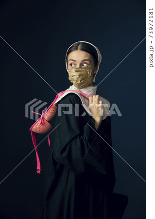 Medieval young woman as a nun in vintage clothing and golden face mask on dark blue background. Concept of comparison of eras, protection from covid Medieval young woman as a nun in vintage clothing and golden face mask on dark blue background. Concept of comparison of eras, protection from covid 73972181