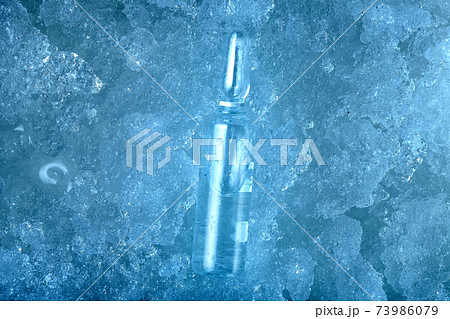 vaccine in glass transparent ampoule with clear liquid with medical preparation for treatment of viral disease is cool to subzero temperatures lying on frozen ice, blue background on pharmaceutical. 73986079