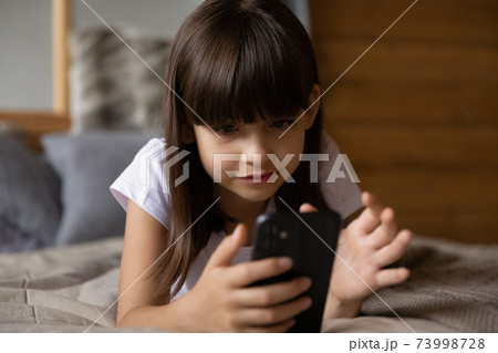 Close up little girl using phone, lying on cozy bed Close up little girl using phone, lying on cozy bed 73998728