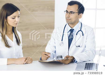 Doctor and patient discussing medical exam results while sitting at the desk in clinic. Male physician using clipboard for filling up medication history record of young woman. Data in medicine 73998919