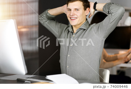 Young businessman with brown curly hair in a green shirt is stretching his hands after working hard on computer in sunny office 74009446