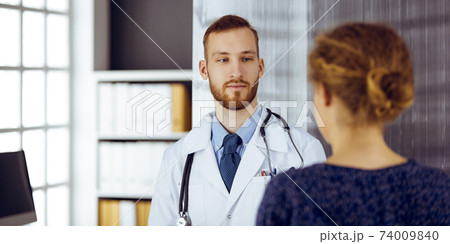 Friendly red-bearded doctor and patient woman discussing current health examination while sitting in sunny clinic. Medicine concept 74009840
