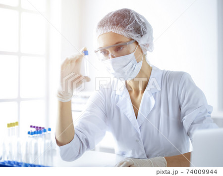 Professional female scientist in protective eyeglasses researching tube with reagents in sunny laboratory. Medicine and science researching Professional female scientist in protective eyeglasses researching tube with reagents in sunny laboratory. Medicine and science researching 74009944