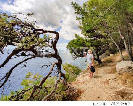 Young active feamle tourist taking a break, drinking water, wearing small backpack while walking on coastal path among pine trees looking for remote cove to swim alone in peace on seaside in Croatia 74010360