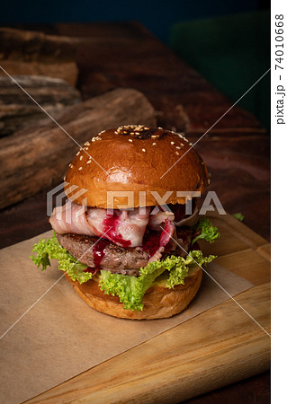 Craft burger with cherry sauce, fried slices of bacon, fresh lettuce next to the decorative wooden lodges. Dish served on a wooden cutting board and wood on background. Street food restaurant concept 74010668