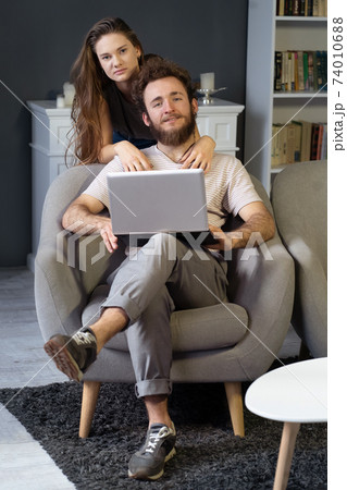 Working together young couple staying quarantined at home with sitting on the arm chairs. Happy young couple sitting on a chair together using laptop. Square cropped Working together young couple staying quarantined at home with sitting on the arm chairs. Happy young couple sitting on a chair together using laptop. Square cropped 74010688