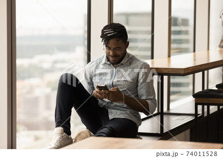 Young african guy sitting on panoramic window sill using smartphone Young african guy sitting on panoramic window sill using smartphone 74011528