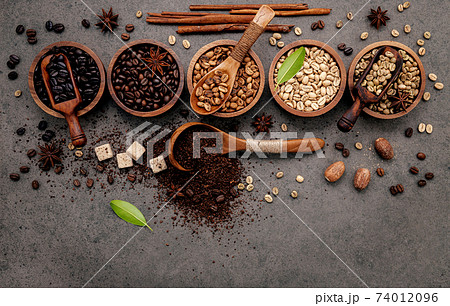 Green and brown unroasted and dark roasted coffee beans in wooden bowl with spoons set up on dark concrete background. 74012096