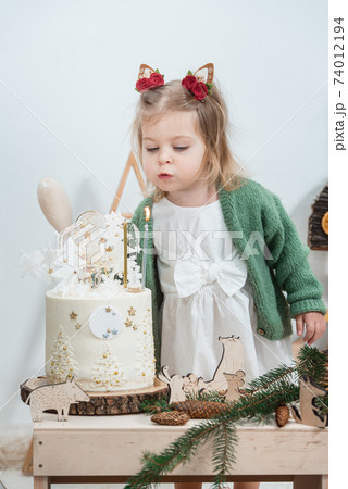Children's winter birthday decorations. Cute little girl in a green cardigan near white birthday cake Children's winter birthday decorations. Cute little girl in a green cardigan near white birthday cake 74012194