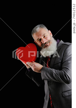 Mature bearded man wear suit holds red heart shaped gift box, isolated black background 74012512