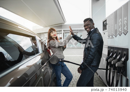 Couple at gas station. Happy joyful multiracial couple of friends or colleagues, posing to camera, having fun while refueling luxury car at petrol station, giving high five each other and smiling 74013940
