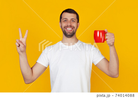 A young man with a beard in a white T-shirt smiles, holds a red cup of coffee and shows a gesture 74014088