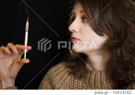 Vaccine concept. Female portrait of caucasian girl holding a syringe and looking at it on black background in studio. Vaccine concept. Female portrait of caucasian girl holding a syringe and looking at it on black background in studio. 74014262