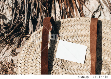 Summer vacation still life. Blank business card mockup on french straw basket with leather handle. Blurred background with rocks and dry palm leaves on Mediterranean Mallorca island. Selective focus. Summer vacation still life. Blank business card mockup on french straw basket with leather handle. Blurred background with rocks and dry palm leaves on Mediterranean Mallorca island. Selective focus. 74016716