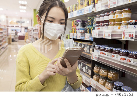 スーパーでスマホを見ながら商品を探すマスク姿女性買い物客 74021552