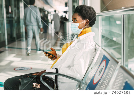 A young African woman in a protective mask sits in a chair in the waiting room and reads emails on her phone. Social distance, pandemic 74027102