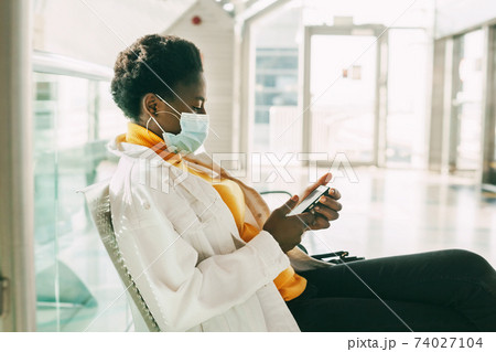 A young African woman in a protective mask sits in a chair in the waiting room and reads emails on her phone. Social distance, pandemic 74027104