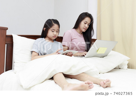 Asian mother and daugther looking laptop and tablet on bed in bedroom. Business women woking with laptop at home with her daugther smiling enjoy with tablet on bed. Work at home concept. 74027365