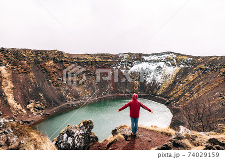 A guy in a red jacket stands on a rock above Lake Kerid - a crater volcanic lake in Iceland. Red volcanic soil, similar to Martian landscapes. Hands thrown upside down and rejoices. 74029159