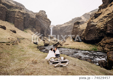 Destination Iceland wedding, near Kvernufoss waterfall. A wedding couple sits on the banks of a mountain river, at a table for a wedding dinner, made of a wooden pallet painted with moss and snags 74029457