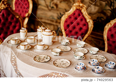 A tea set with a blue pattern and a golden set on a tray on a white mantel with molding against the background of a red wall with patterns. A tea set with a blue pattern and a golden set on a tray on a white mantel with molding against the background of a red wall with patterns. 74029563