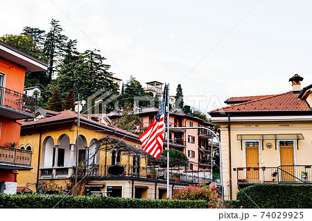 The flag of the United States of America on a flagpole in the garden near residential buildings among the trees. 74029925