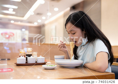 Asian child girl in a face mask under the chin,eating food,plastic shield partition on the tables to protect during new outbreak of COVID-19,safe distance,keeping a social distancing,new normal life 74031426