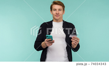 Cheerful sports red-haired guy conducts online payment and looks at the smartphone screen on a blue background. Cheerful sports red-haired guy conducts online payment and looks at the smartphone screen on a blue background. 74034343