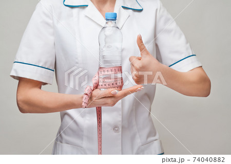 Portrait of young caucasian woman health care professional holding bottle of water and measuring tape standing over light grey background Portrait of young caucasian woman health care professional holding bottle of water and measuring tape standing over light grey background 74040882