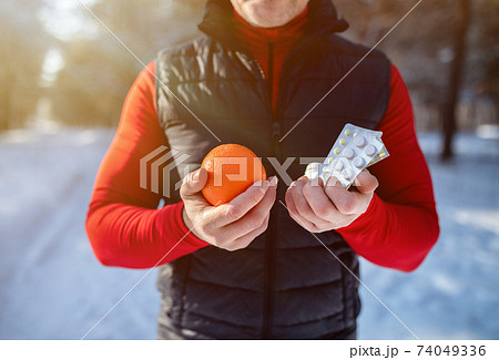 Healthy lifestyle vs medical treatment. Closeup of senior sportsman holding orange and pills at snowy winter park Healthy lifestyle vs medical treatment. Closeup of senior sportsman holding orange and pills at snowy winter park 74049336