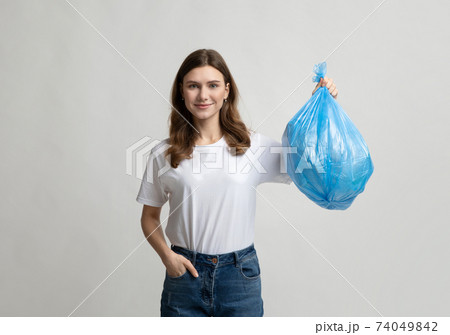 Happy young woman holding blue plastic garbage bag, standing on grey background Happy young woman holding blue plastic garbage bag, standing on grey background 74049842