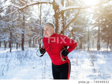 Functional training. Portrait of senior man doing exercises with TRX fitness straps at snowy park in winter Functional training. Portrait of senior man doing exercises with TRX fitness straps at snowy park in winter 74049932