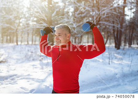 Senior man exercising with dumbbells outdoors in winter. Mature bodybuilder working out his biceps at snowy park Senior man exercising with dumbbells outdoors in winter. Mature bodybuilder working out his biceps at snowy park 74050078