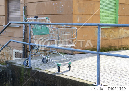 Abandoned empty grocery cart in an old street courtyard with shabby walls and closed shutters. Discarded shopping cart chained to the handrails of a concrete staircase Abandoned empty grocery cart in an old street courtyard with shabby walls and closed shutters. Discarded shopping cart chained to the handrails of a concrete staircase 74051569