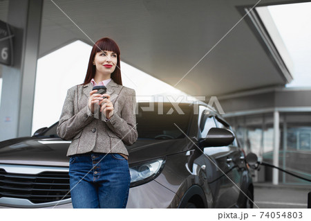 Transport, people, coffee break concept. Pretty young Caucasian girl in casual clothes, leaning on the car and enjoying her coffee to go, while refueling auto at petrol station Transport, people, coffee break concept. Pretty young Caucasian girl in casual clothes, leaning on the car and enjoying her coffee to go, while refueling auto at petrol station 74054803