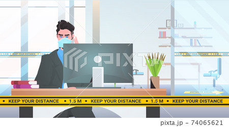 businessman in mask sitting at workplace desk with yellow tape for social distancing 74065621