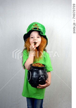 Nice littel girl with decorative red beard in green dress and leprechaun's hat, kissing golden coin with four-petal lucky shamrock leaf, holds cast iron pot, full of golden treasure. St. Patrick's Day Nice littel girl with decorative red beard in green dress and leprechaun's hat, kissing golden coin with four-petal lucky shamrock leaf, holds cast iron pot, full of golden treasure. St. Patrick's Day 74070709