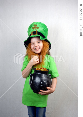 St. Patrick's Day. Charming seven years old girl laugh and showing off gold coin with four-petal lucky clover leaf and  cast iron pot, full of leprechaun golden treasure 74070710