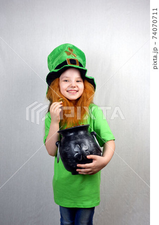Saint Patricks Day. Cute laughting small girl with decorative red beard in green dress and leprechaun's hat, holds cast iron pot, full of golden treasure, showing off golden coin with four-petal 74070711